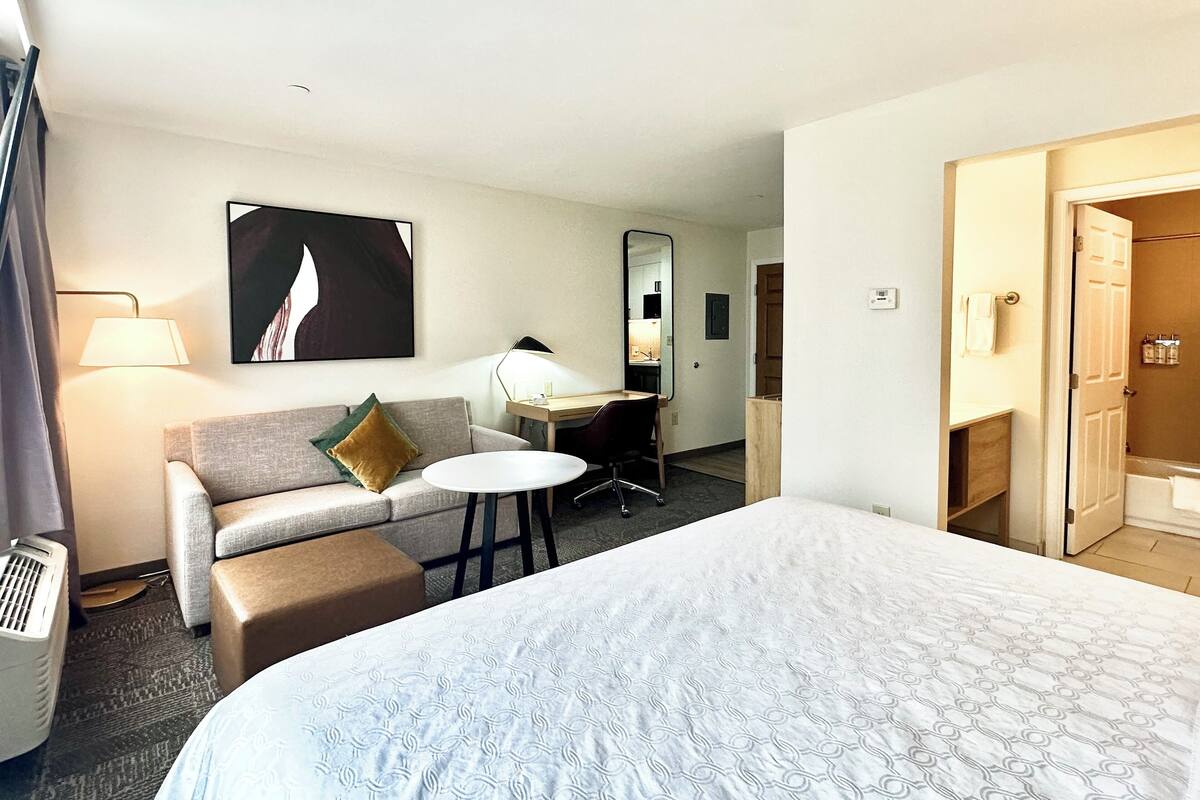 A hotel room with a large bed, a sofa, a small table, a desk, a mirror, a lamp, and a bathroom visible through an open door.