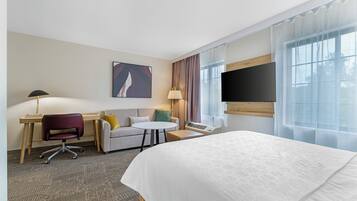Une chambre d’hôtel comprenant un lit, un bureau, une chaise, un canapé, deux lampes, une télévision et une fenêtre avec des rideaux.