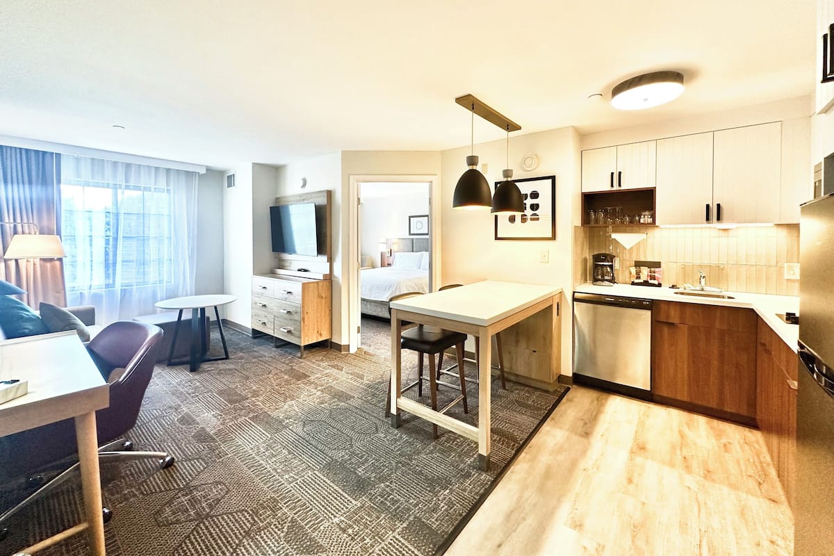 A modern hotel room with a dining area, kitchenette, and a bed.