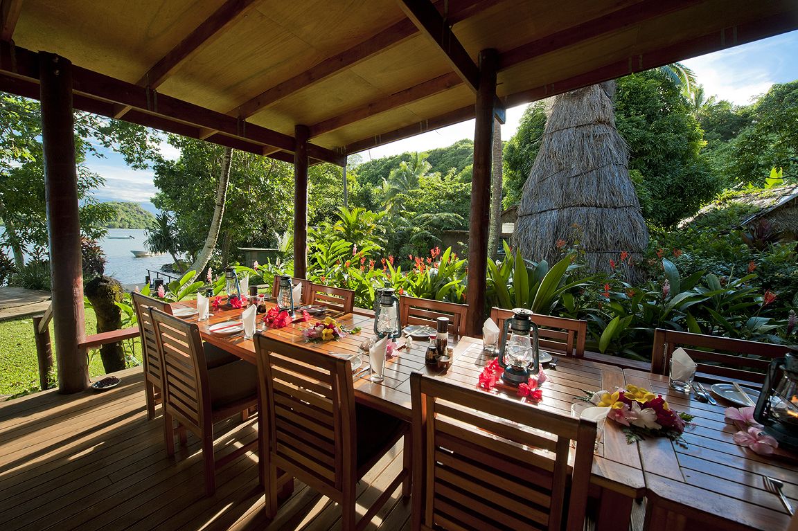 Eine Holzdielen-Terrasse mit Esstisch, umgeben von üppiger Vegetation und Blick auf ein Gewässer.