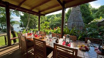 Eine Holzdielen-Terrasse mit Esstisch, umgeben von üppiger Vegetation und Blick auf ein Gewässer.
