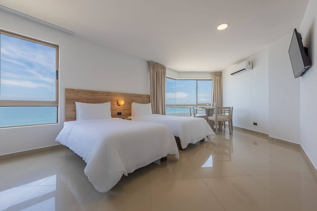 A hotel room with two beds, a large window with a view, a TV mounted on the wall, and an air conditioning unit.
