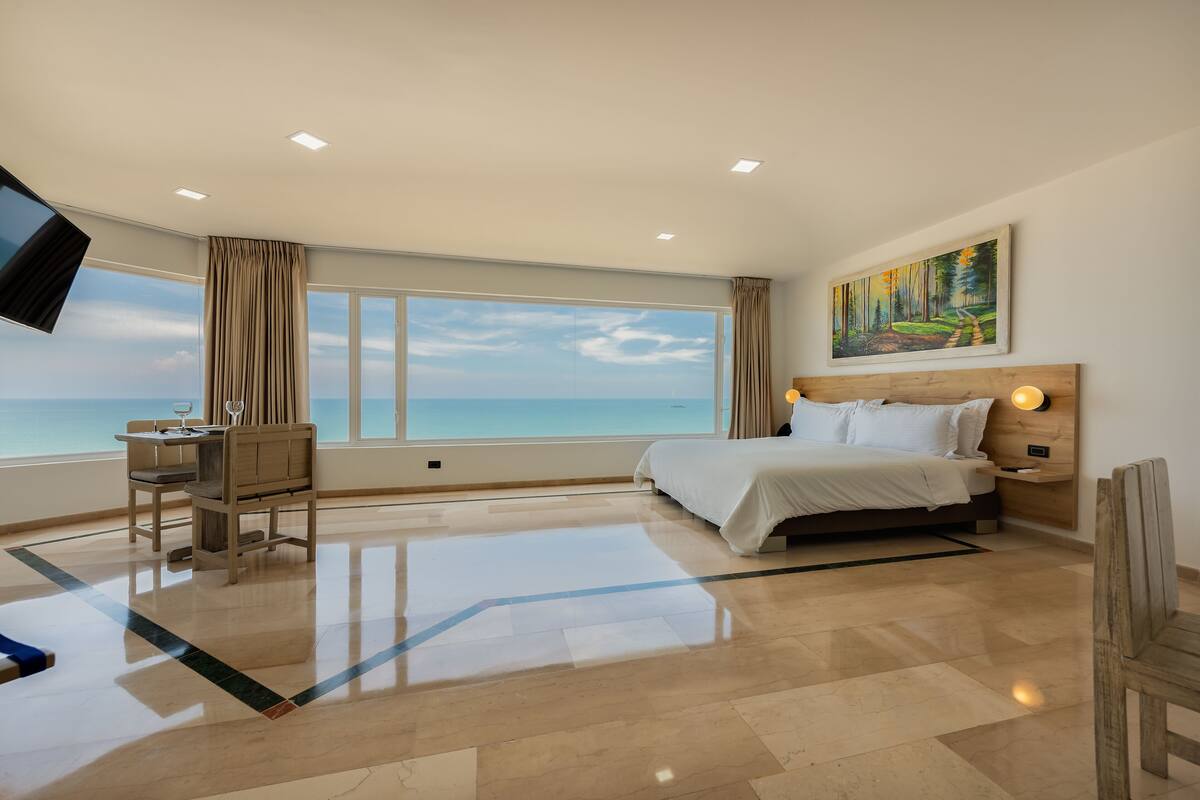 A hotel room with a large window, a bed, a desk with a chair, and a painting on the wall.