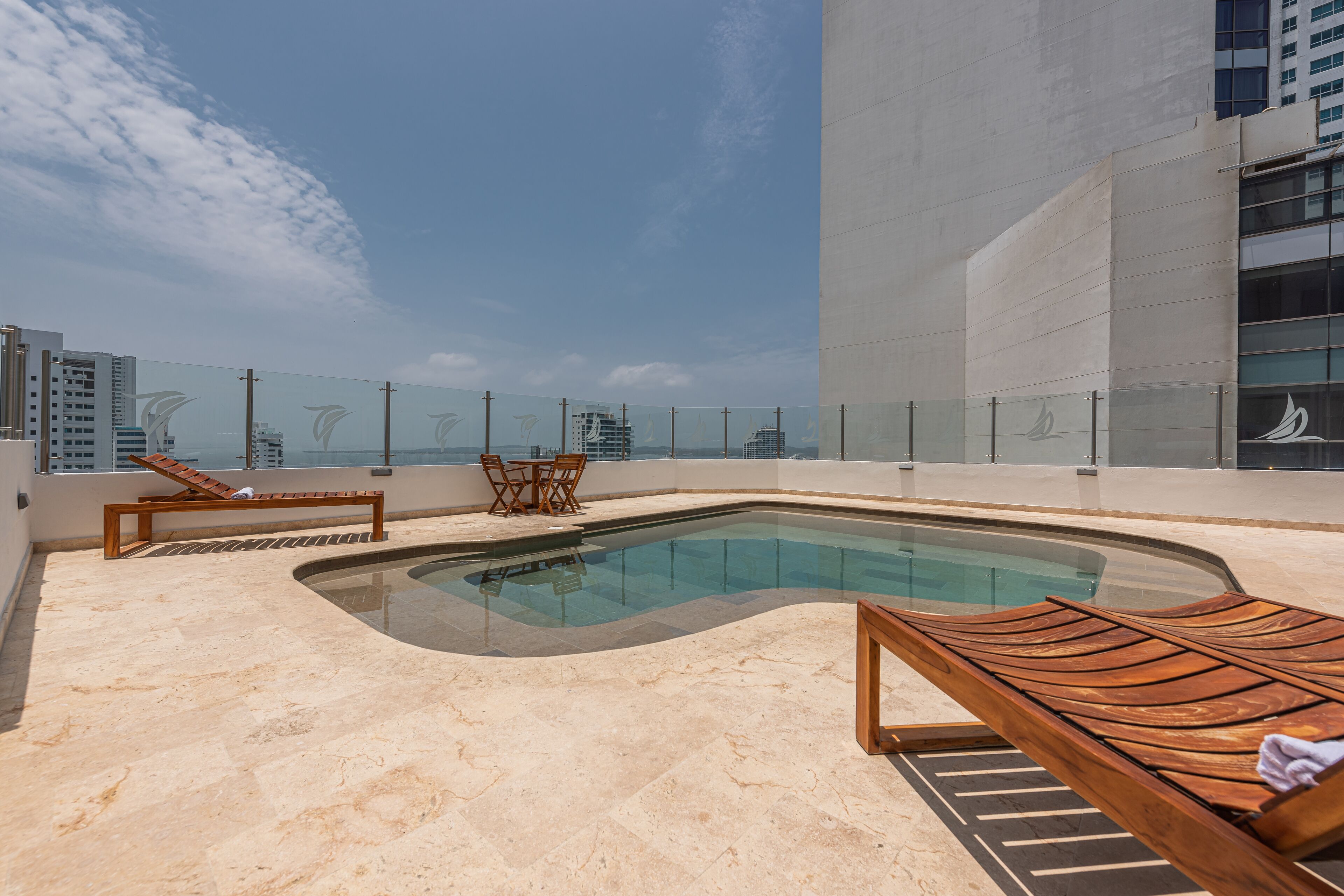 A rooftop pool area with a glass barrier, lounge chairs, and a small table.