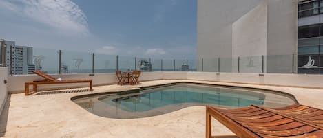 A rooftop pool area with a glass barrier, lounge chairs, and a small table.