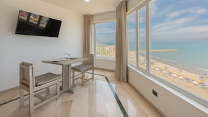 Una habitación con una televisión montada en la pared, una pequeña mesa con sillas y un amplio ventanal con vistas a la playa.