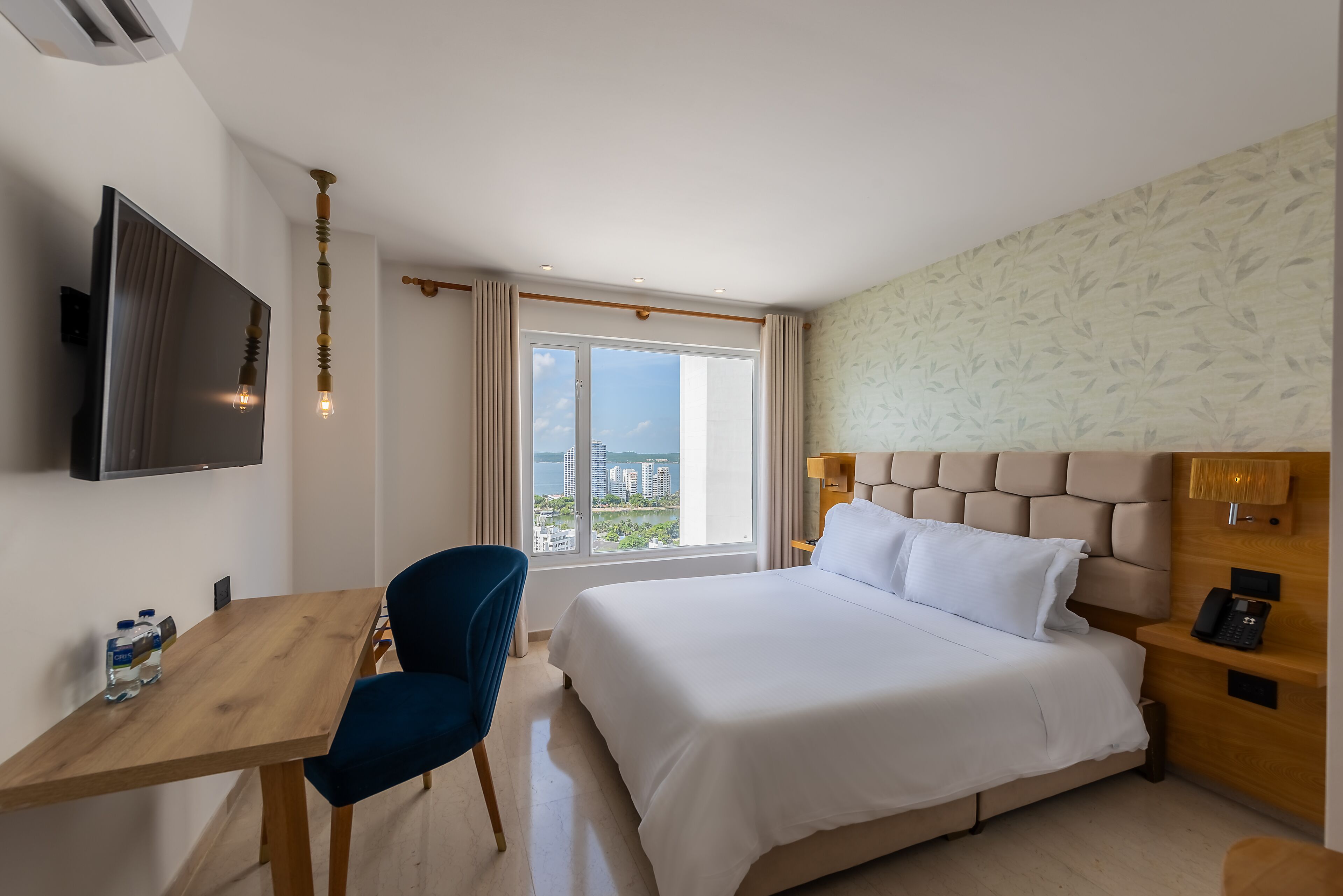 Hotelkamer met een bed, een bureau, een stoel, een televisie en een raam met uitzicht op gebouwen en groen.