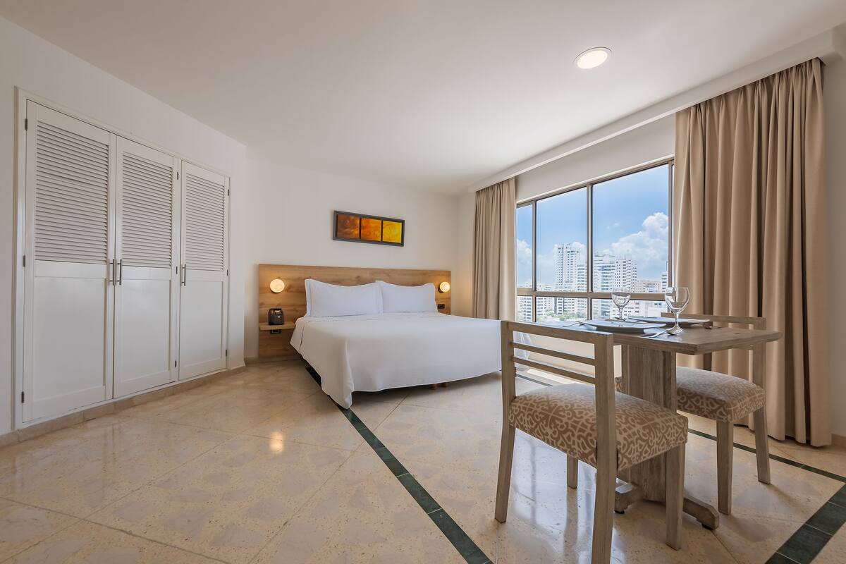 A hotel room with a bed, a desk with a chair, a window with a city view, and a wardrobe.