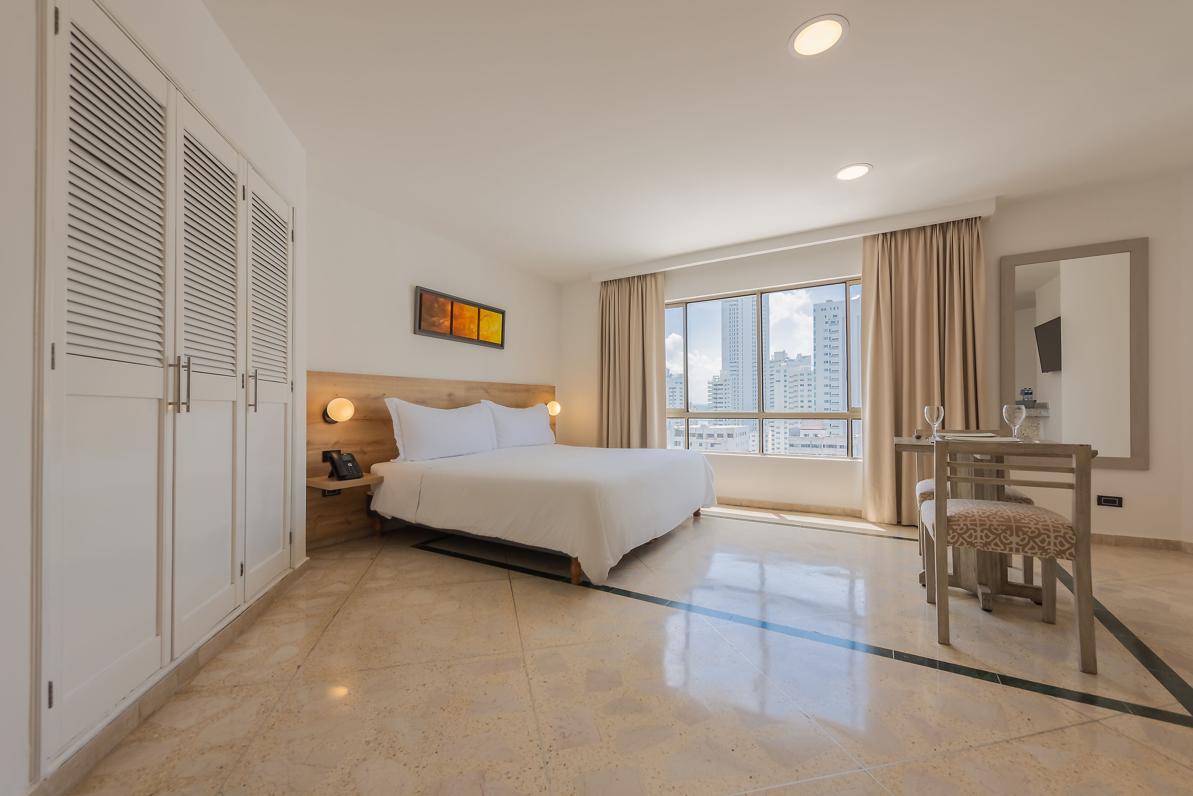 A hotel room with a bed, bedside table, lamp, and a view of the city through large windows.