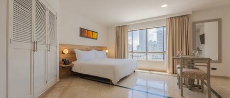 Habitación de hotel con cama, mesita de noche, lámpara y vista a la ciudad a través de amplias ventanas.