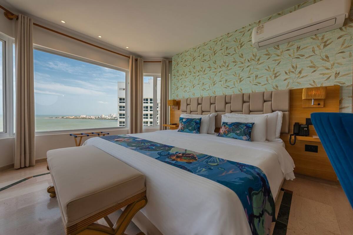 A hotel room with a large bed, a view of the sea, a wooden headboard, and a nightstand with a phone.