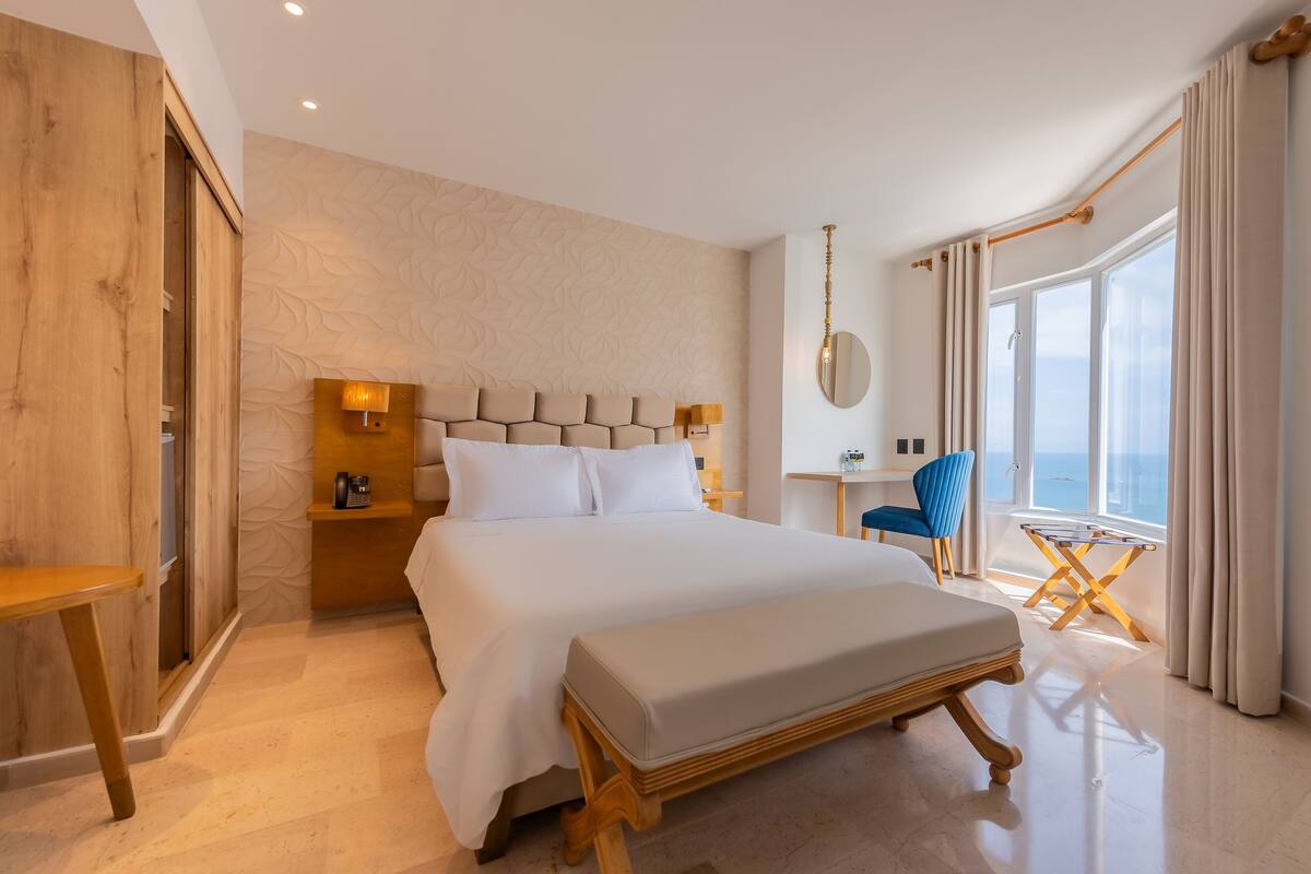 A modern hotel room with a large bed, a wooden bench, a small table, and a view of the sea through large windows.