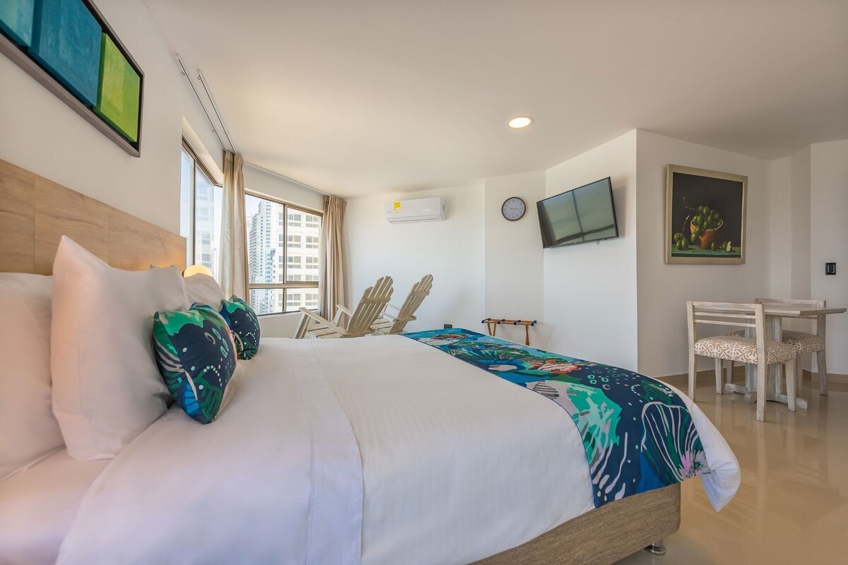 A hotel room with a bed, two chairs, a desk, a TV, and a painting on the wall.