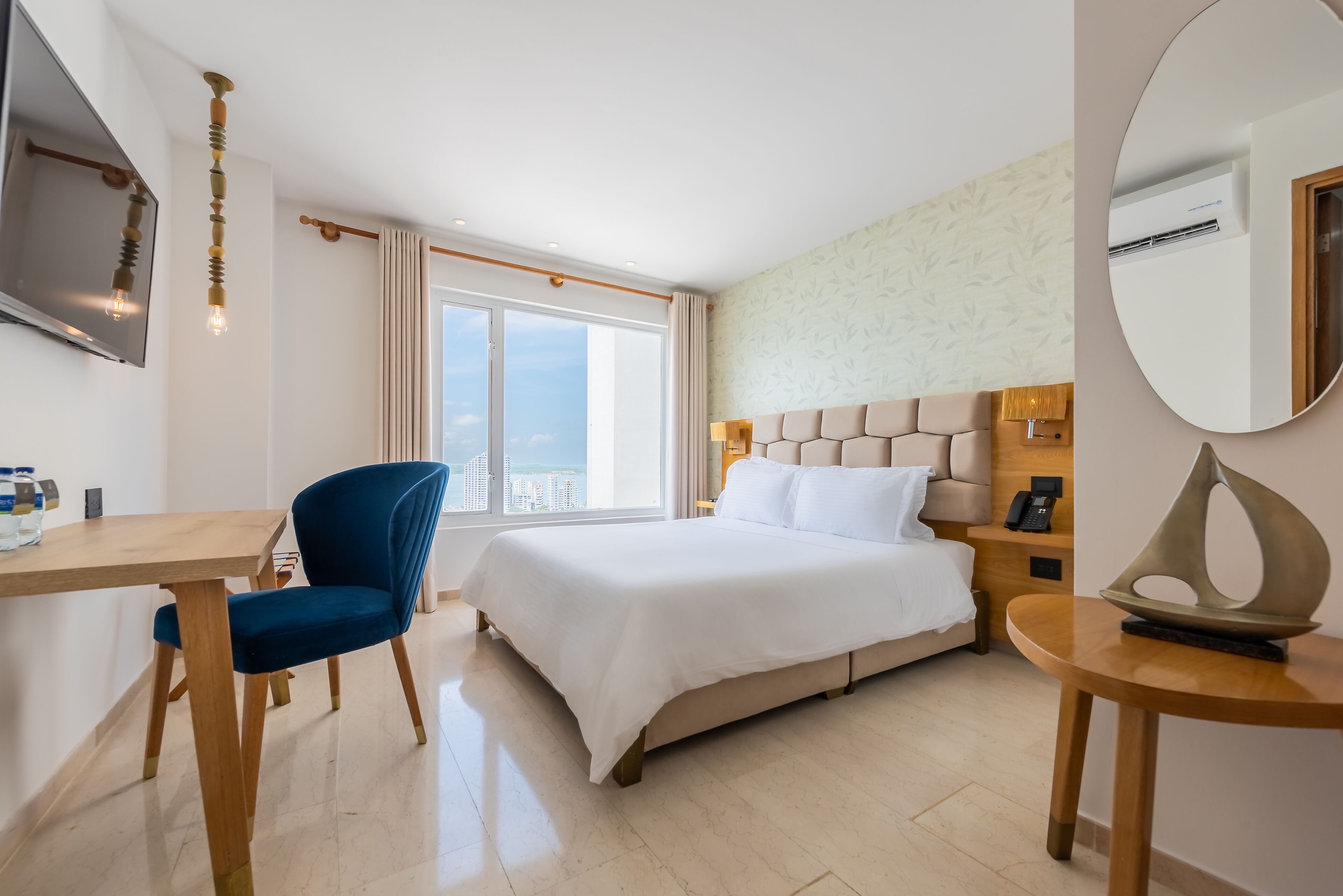 A hotel room with a bed, a desk, a chair, a bedside table, and a large window with a view.