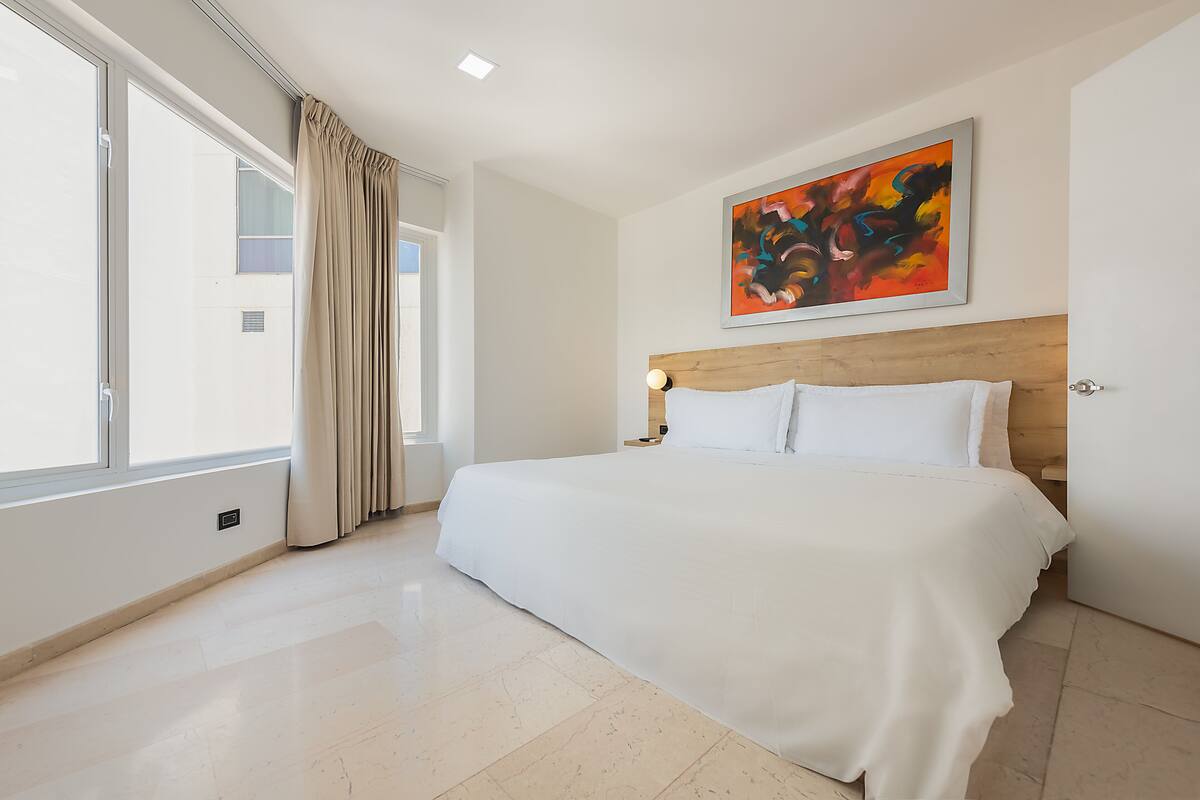 A modern hotel room with a large bed, a wooden headboard, a large window with curtains, and a colorful abstract painting on the wall.