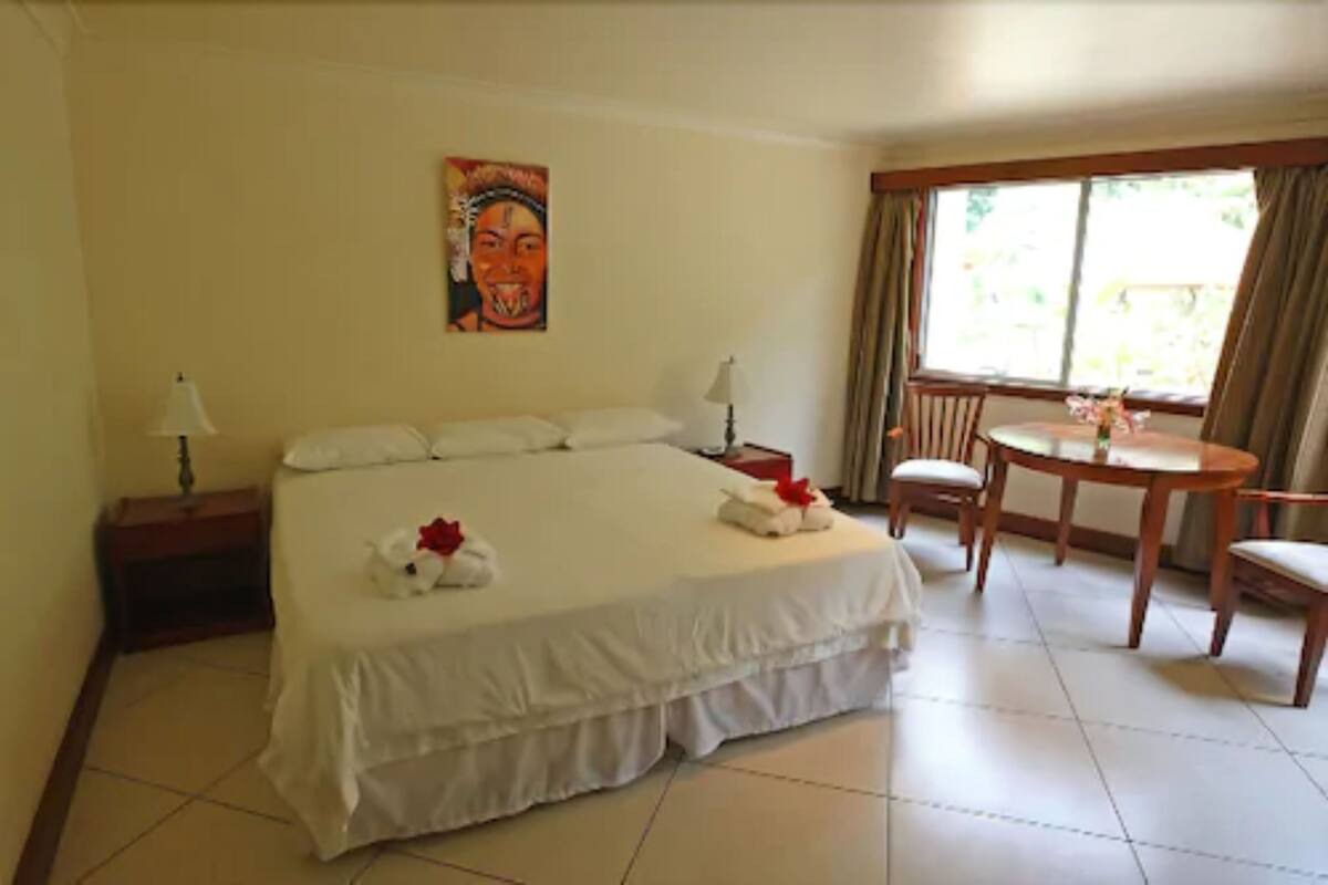 Une chambre d’hôtel avec un grand lit, une petite table avec des chaises, un tableau au mur et une fenêtre avec des rideaux.