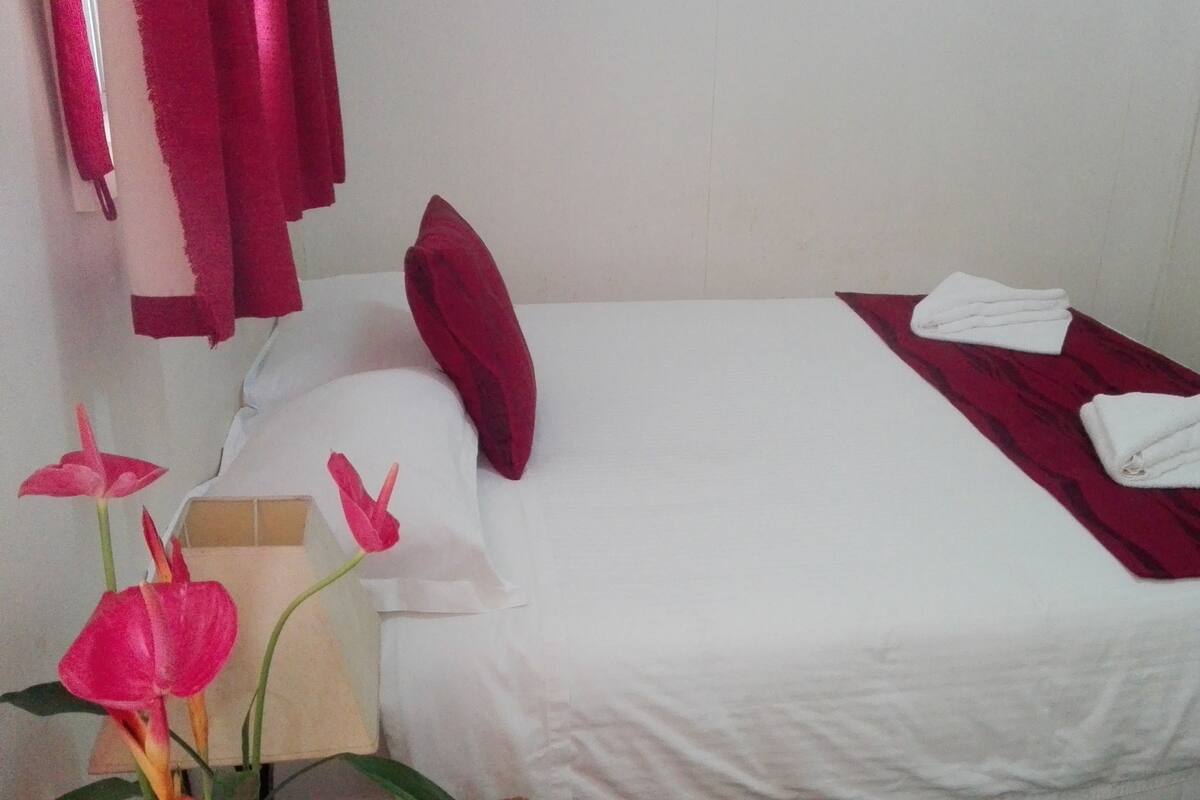 Une chambre d’hôtel avec un lit simple, du linge de lit blanc, un oreiller rouge et une couvre-lit rouge. Il y a des serviettes blanches sur le lit.