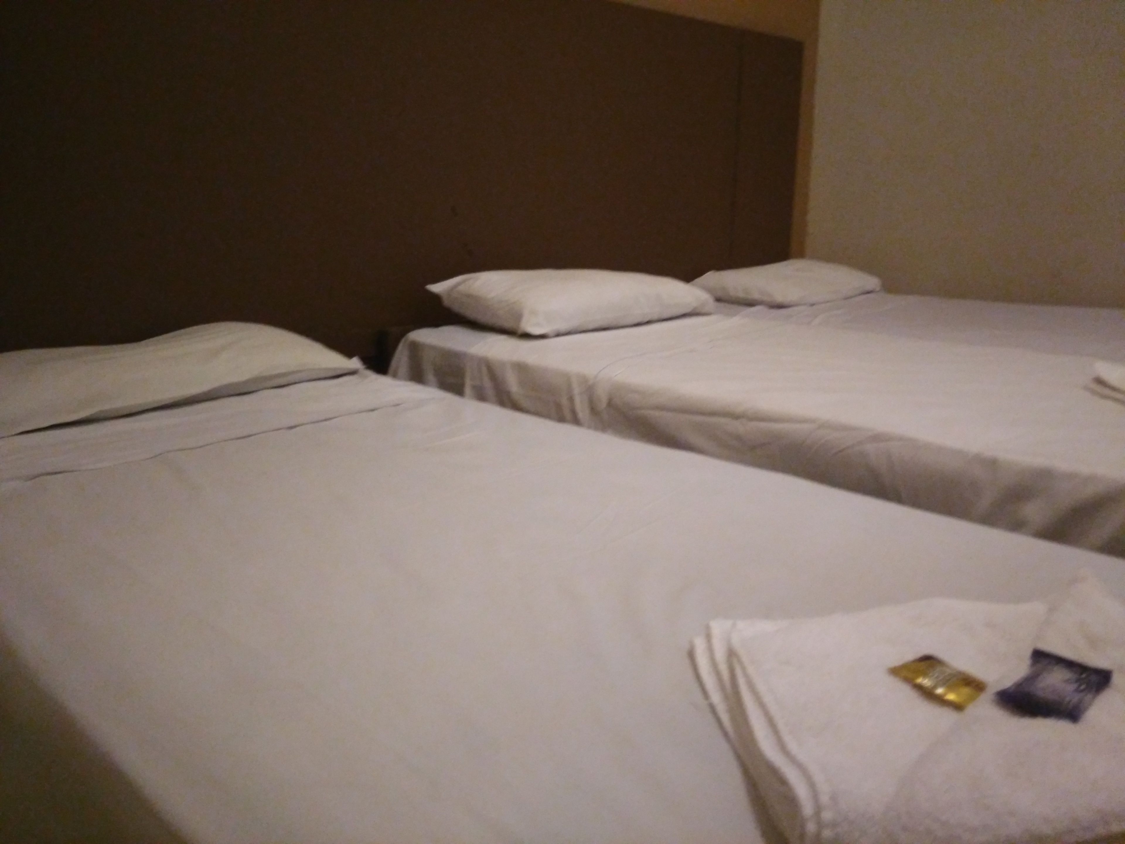 Two single beds with white linens and pillows, a brown headboard, and a small table with a towel and two packets.