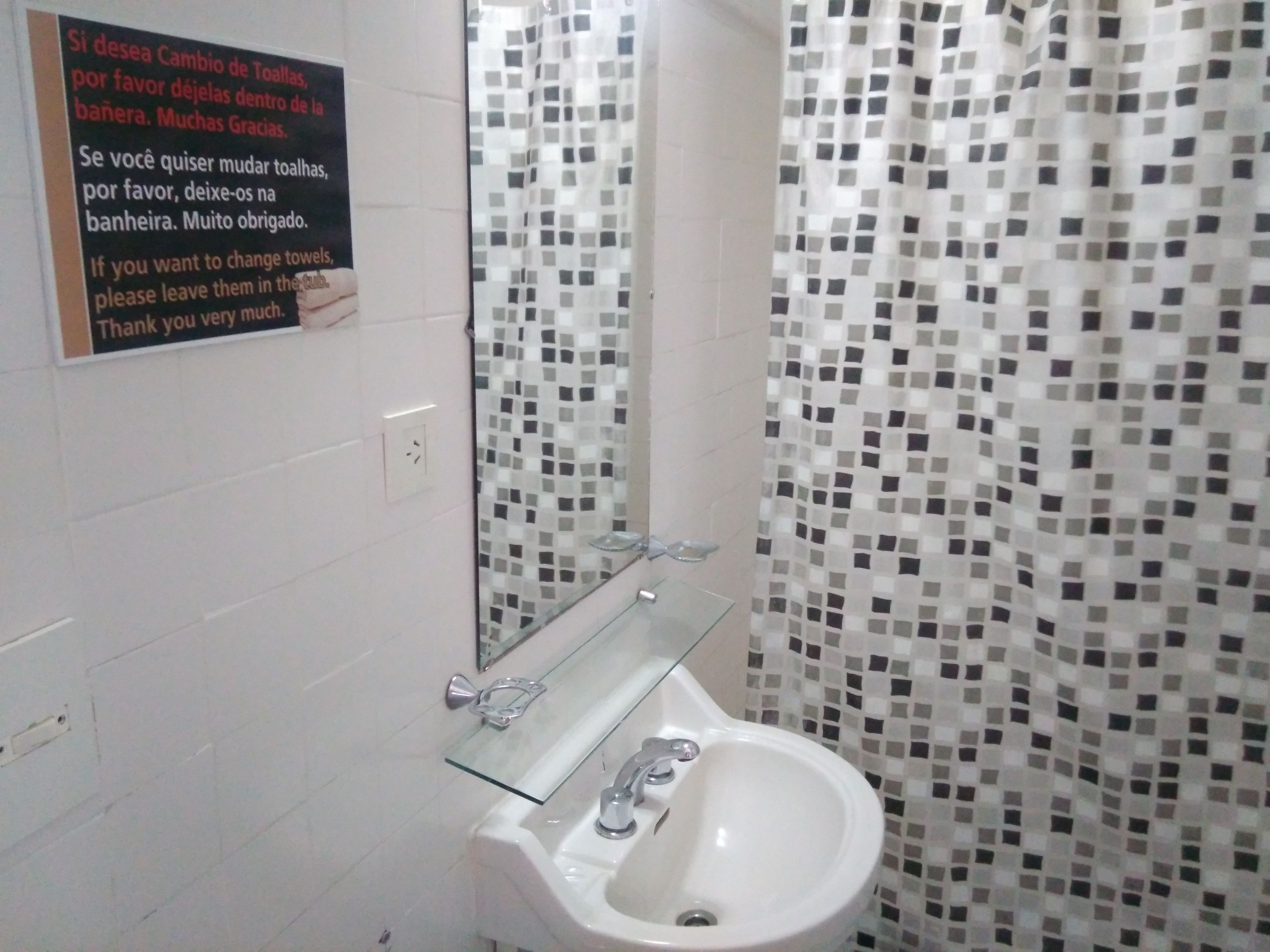 Ein Badezimmer mit einem weißen Waschbecken, einer Glasablage und einem Spiegel. An der Wand befindet sich ein Schild mit spanischem Text. Der Duschvorhang hat ein Mosaikmuster.