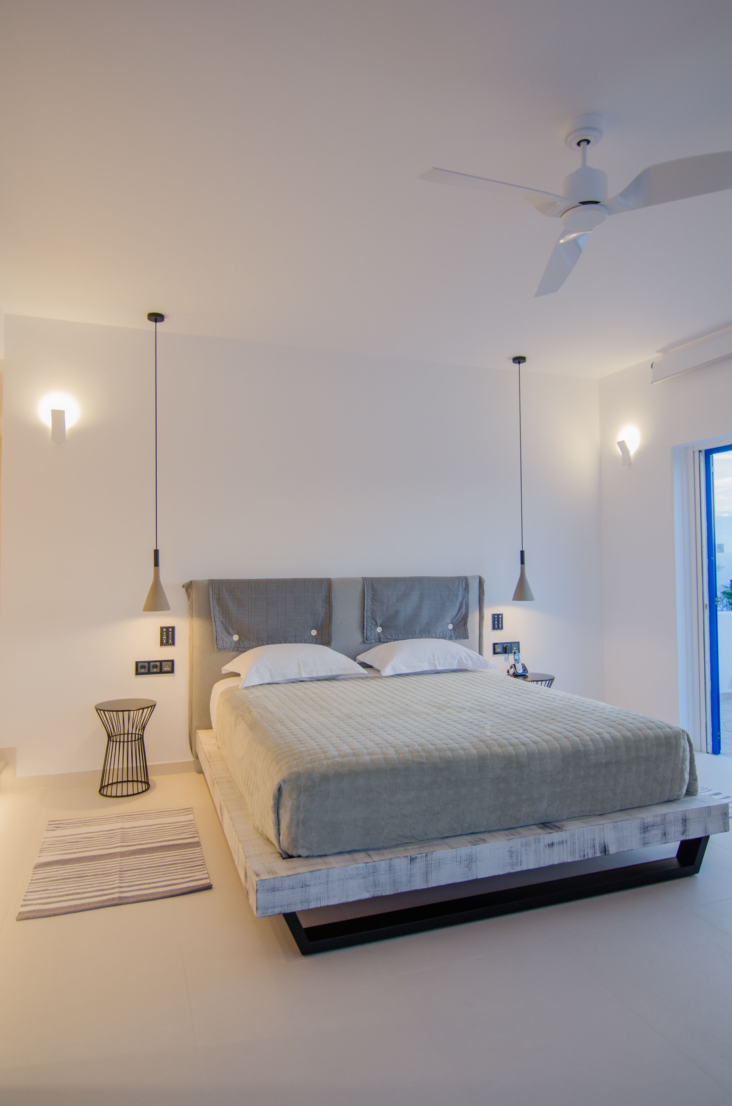 A modern bedroom with a large bed, two pendant lights, a ceiling fan, and a view of the outdoors.