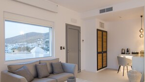 A modern living room with a sofa, coffee table, and a view of a town through a large window. - Lofos Village Hotel (Ios)