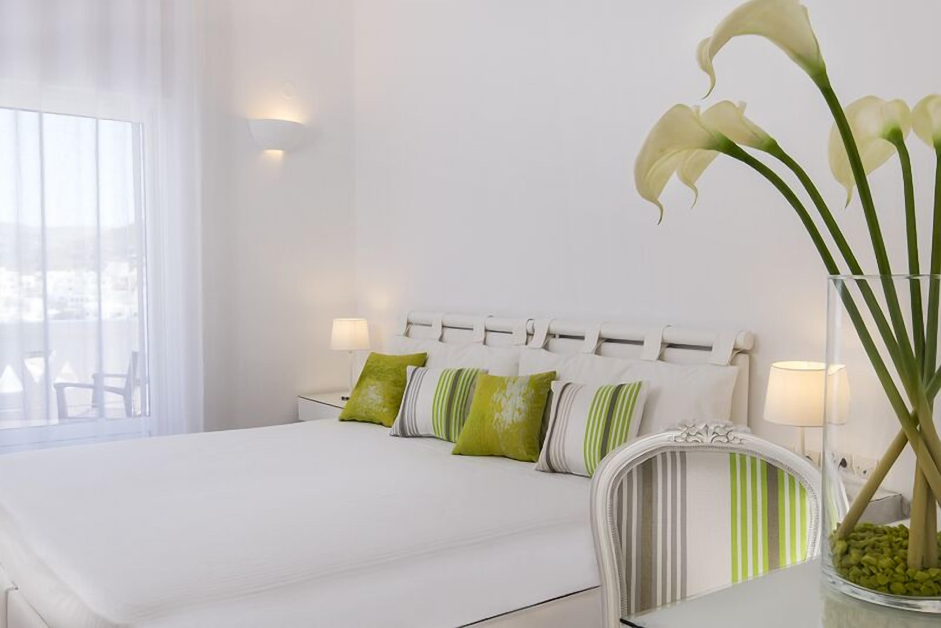 A neatly made bed with white bedding and green and white striped pillows, a white headboard, and a vase with white flowers on a bedside table.