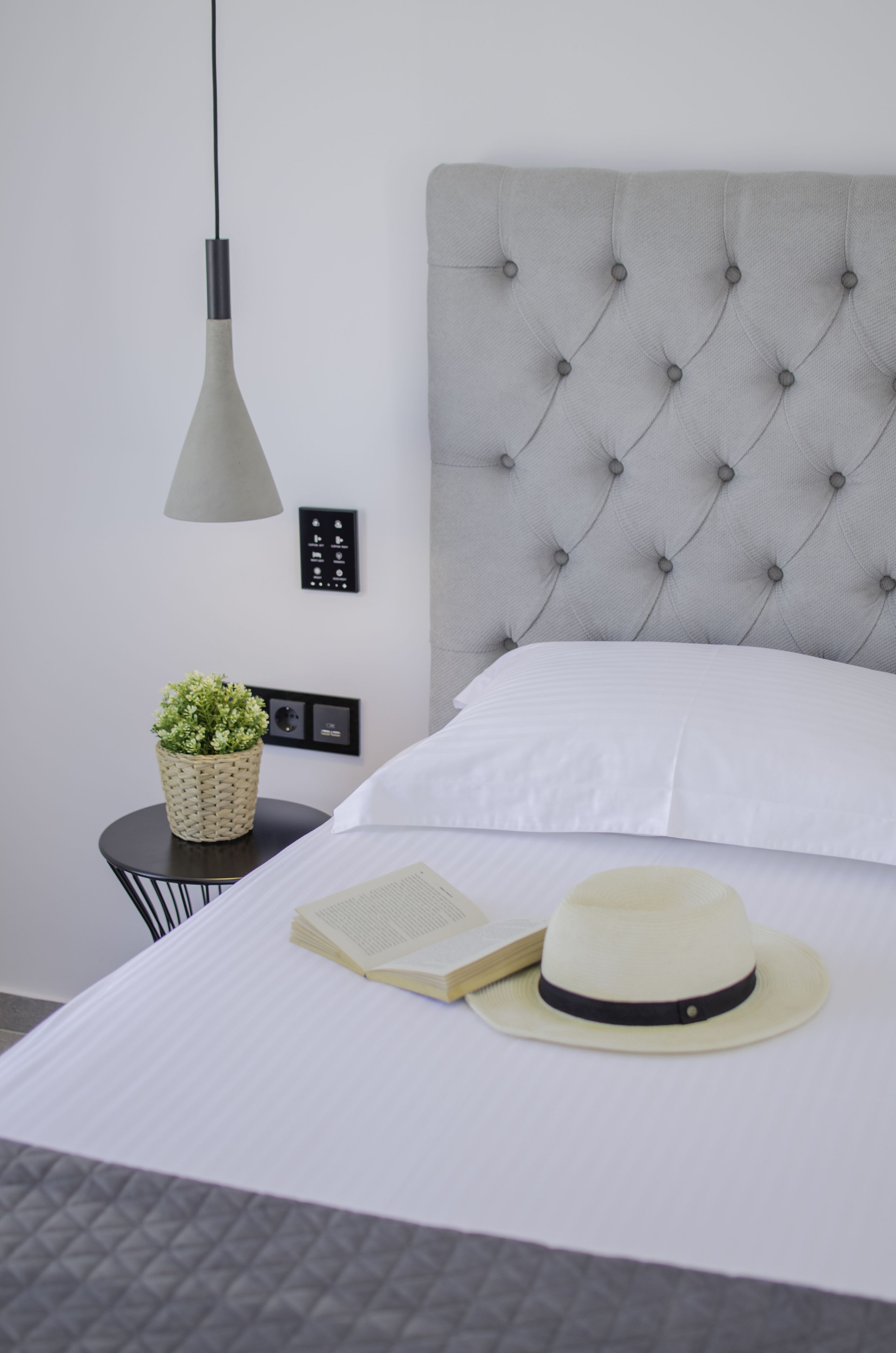 Een net opgemaakt bed met een grijs hoofdbord, een bedlampje, een kleine bloempot, een open boek en een hoed op het bed.