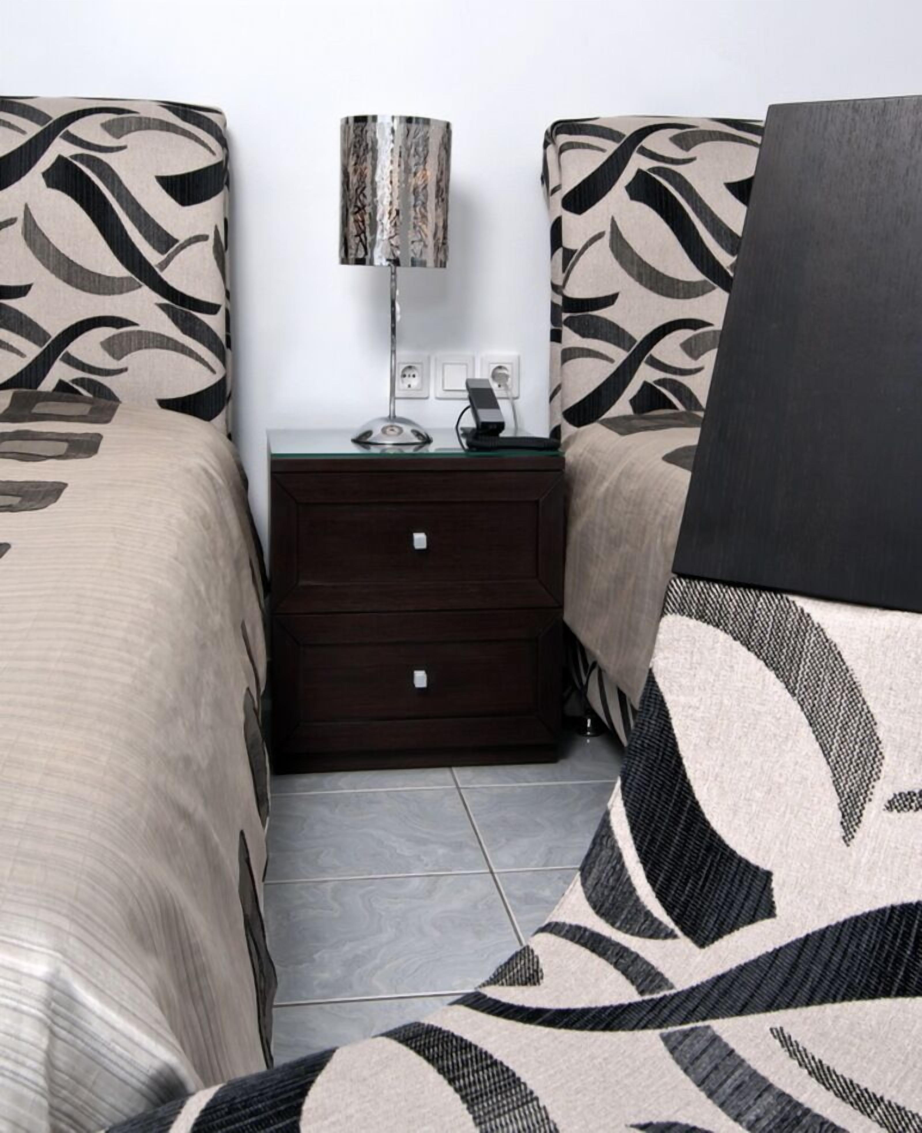 A hotel room with two beds, a nightstand with a lamp, and a small drawer unit.