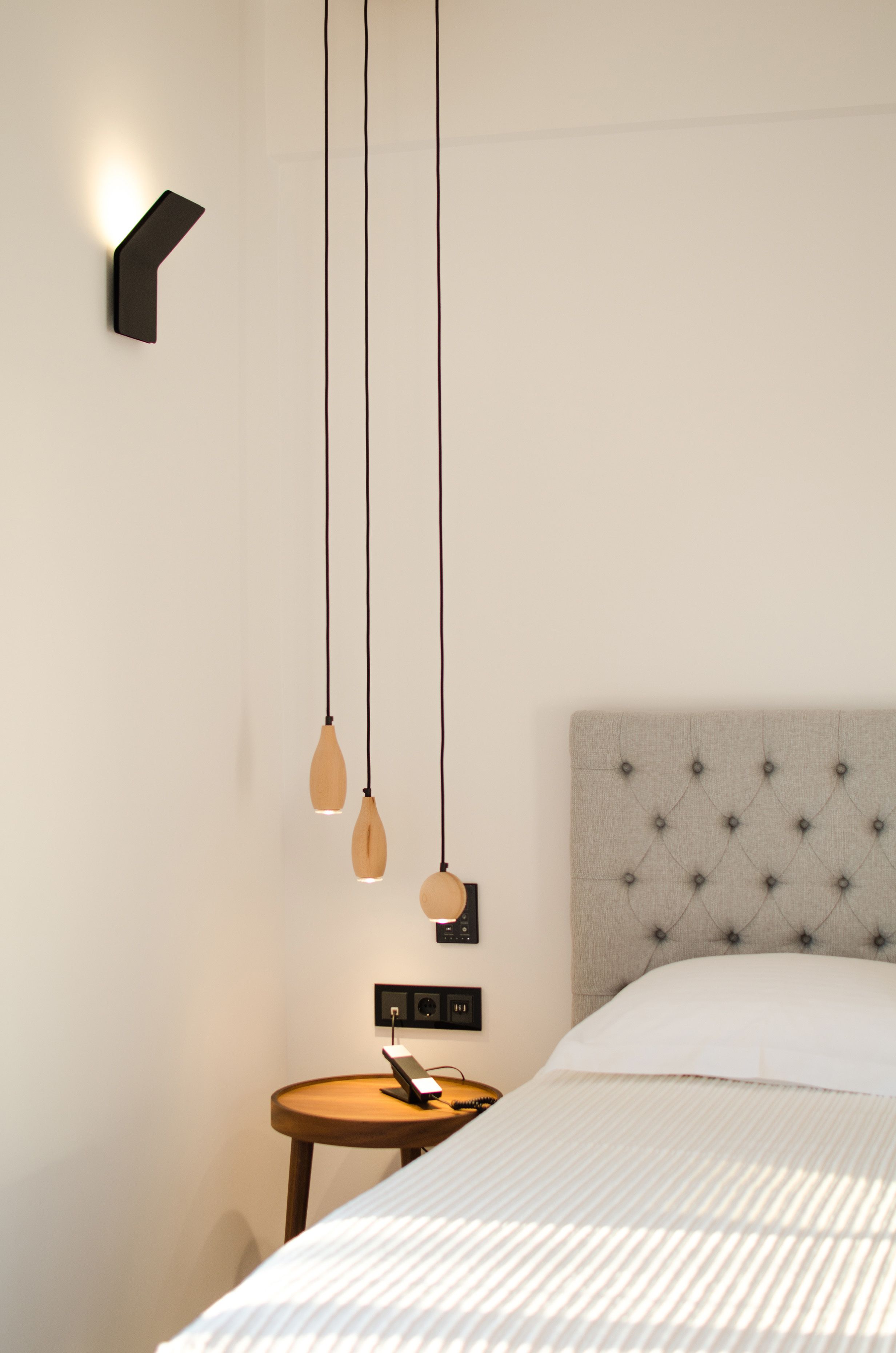 A hotel room with a bed, a nightstand, a wall-mounted light, and hanging pendant lights.