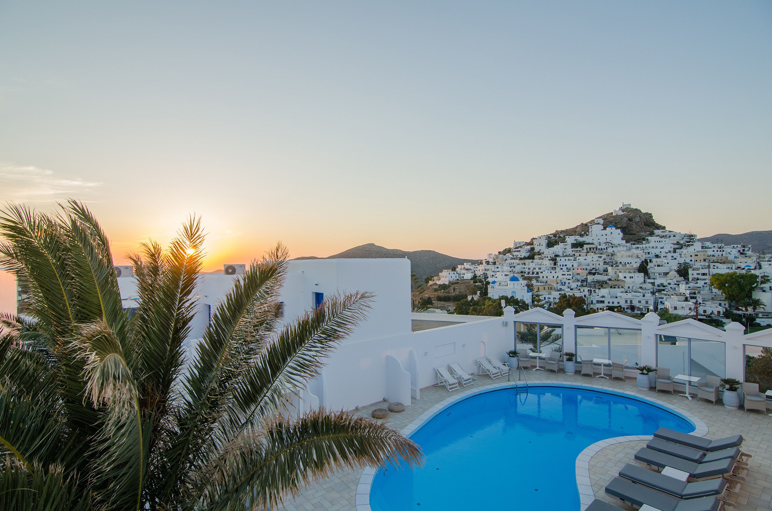 A poolside area with lounge chairs, a clear blue pool, and a view of a white-washed village with a hill in the background.