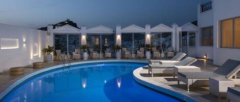 A modern outdoor swimming pool with lounge chairs and sun loungers, surrounded by white walls and large windows.