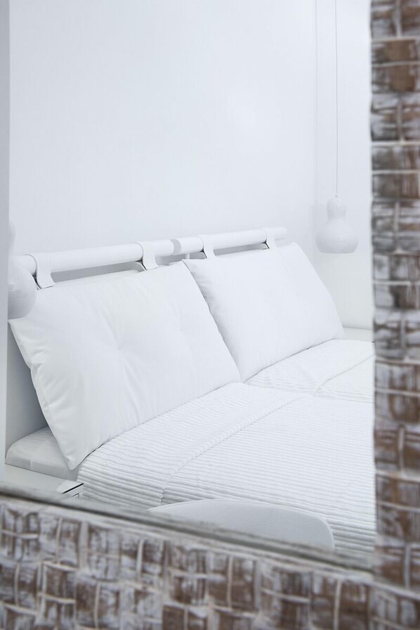 A white bed with a headboard, white bedding, and a brick-patterned wall. - Lofos Village Hotel (Ios)