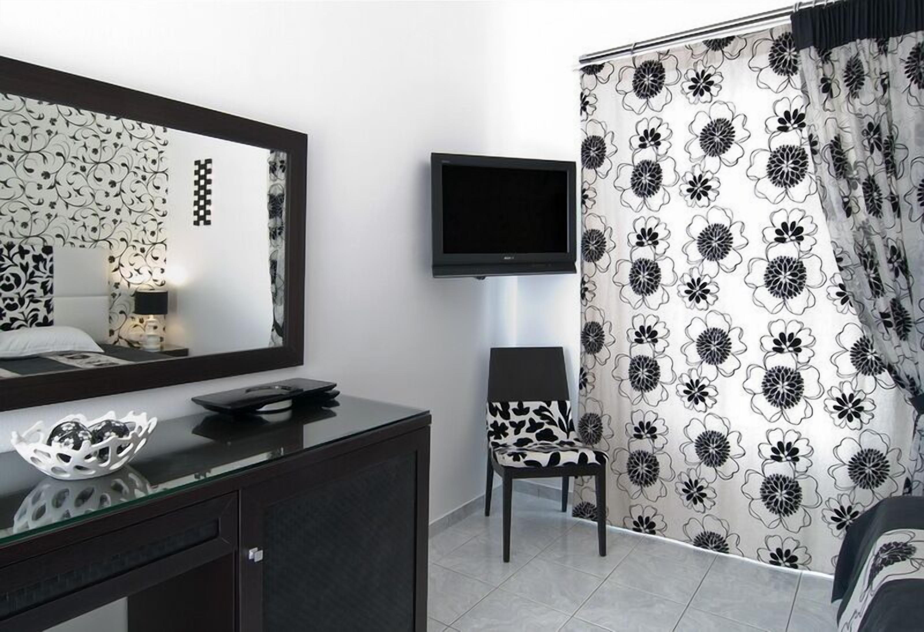 A modern bedroom with a dark wooden dresser, a flat-screen TV mounted on the wall, a patterned chair, and a bed with a white and black floral curtain.