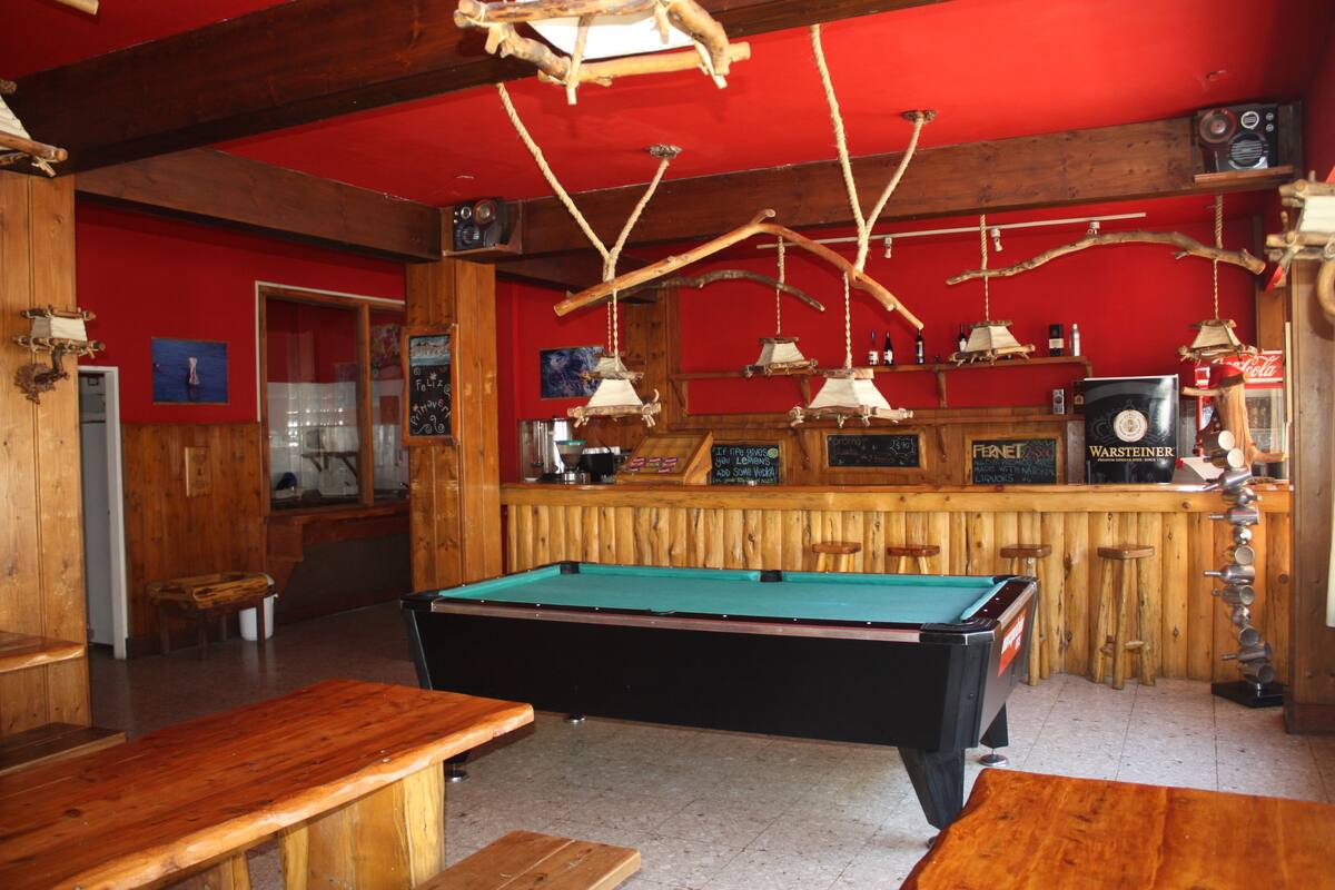 A pool table in a room with wooden walls and a red ceiling. There are wooden benches and a bar area with bottles and a sign.