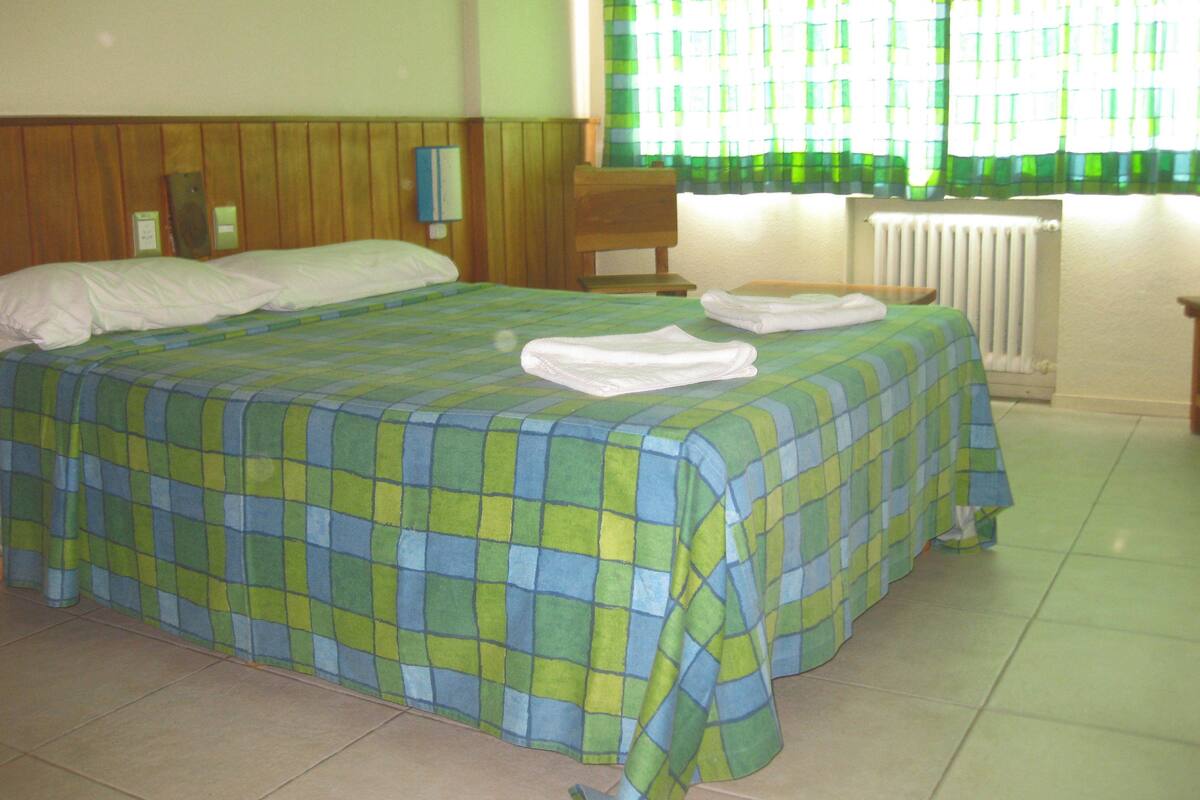 A bed with a checkered bedspread, a wooden headboard, a nightstand with a lamp, and a window with green curtains.