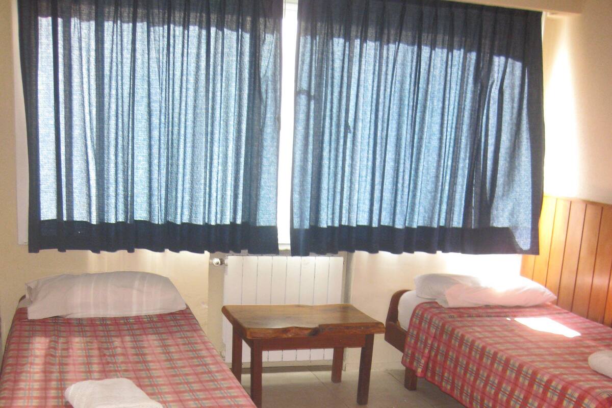A room with two single beds, each with a plaid bedspread and a rolled towel. There is a wooden table between the beds.