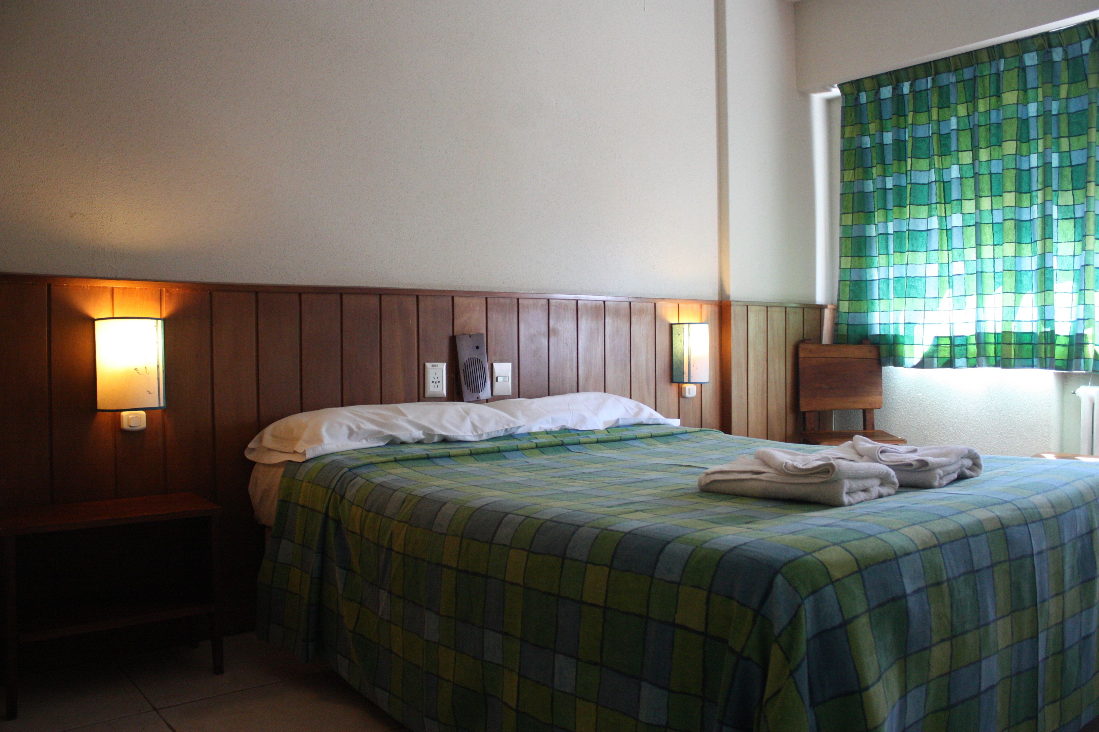 Een hotelkamer met een houten hoofdeinde, een bed met een groen geruit sprei, twee bedlampen en een raam met een groen geruit gordijn.