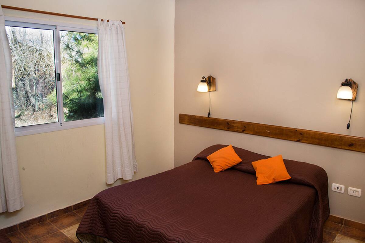 A bedroom with a bed, two wall-mounted lights, a window with curtains, and a view of trees outside.