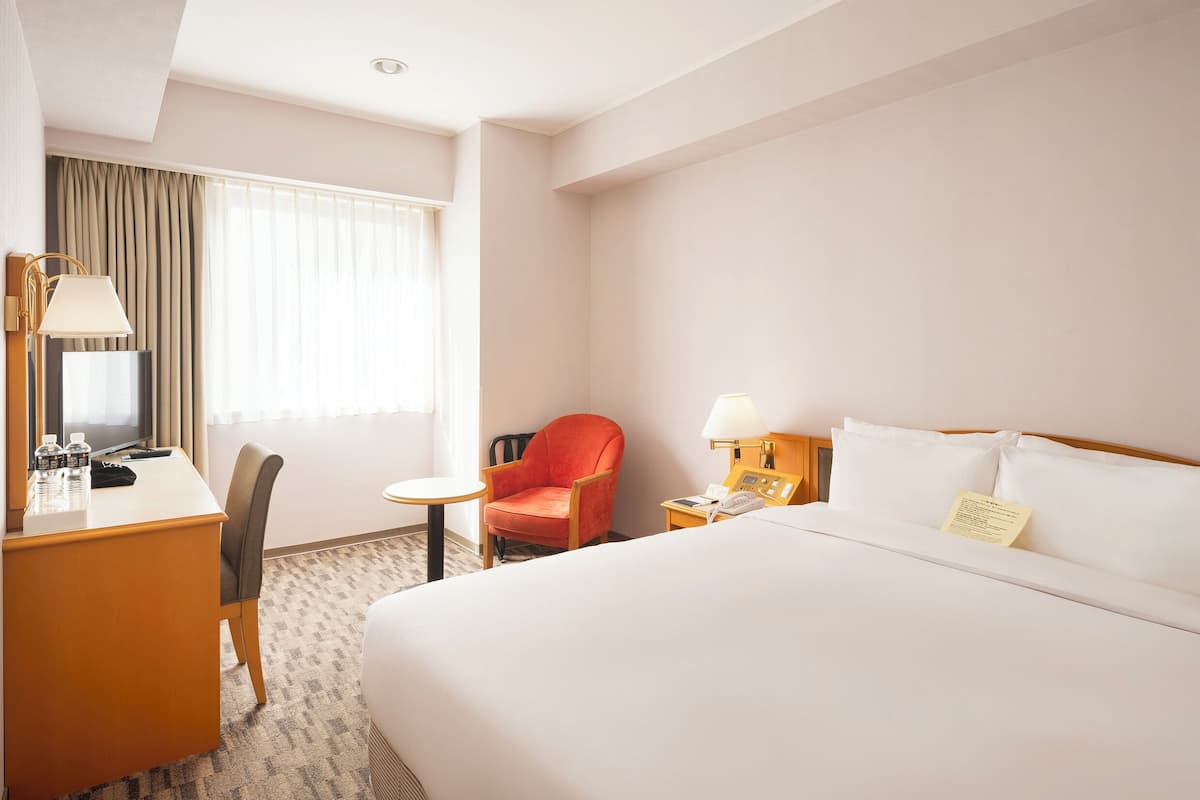A hotel room with a bed, a desk, a chair, a small table, a lamp, a TV, and curtains.