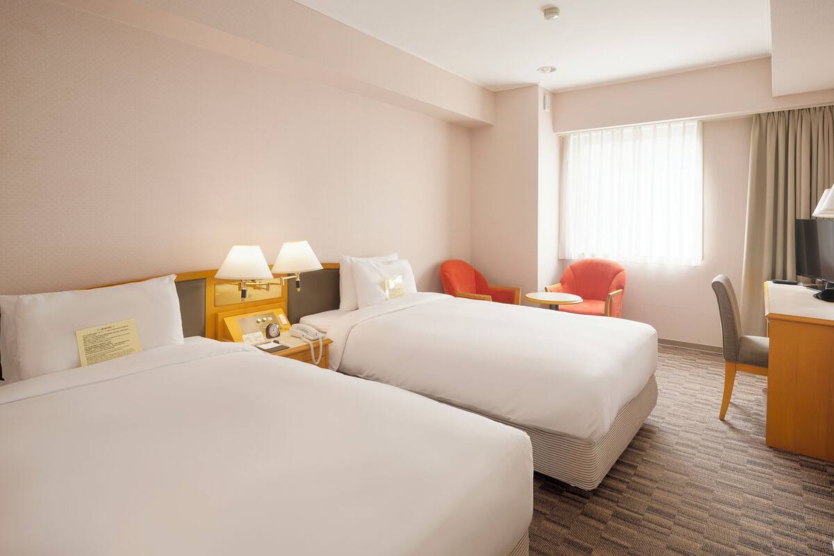 A hotel room with two beds, a desk, a chair, a small table, and a window with curtains.