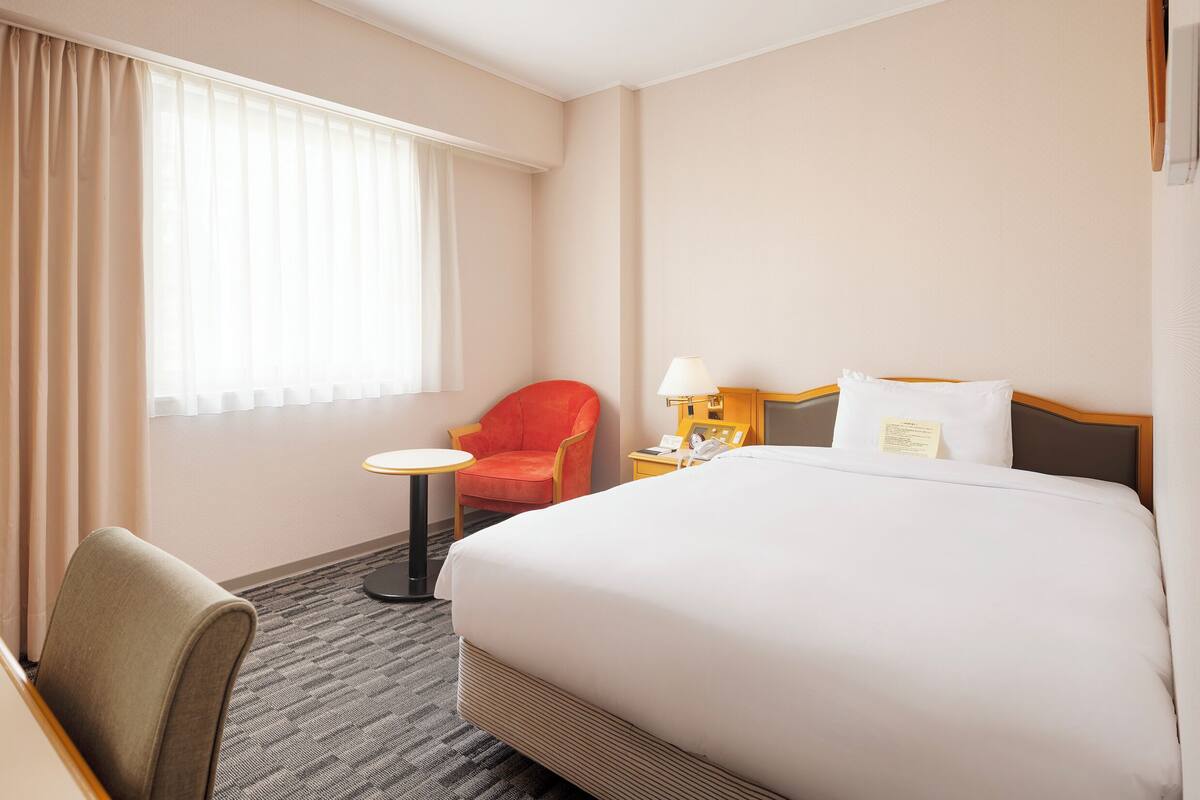 A hotel room with a bed, a chair, a small table, and a nightstand with a lamp.