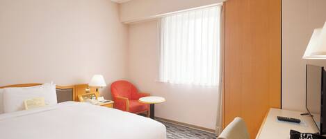 A hotel room with a bed, a desk, a chair, a small table, a lamp, a remote control, and a window with curtains.
