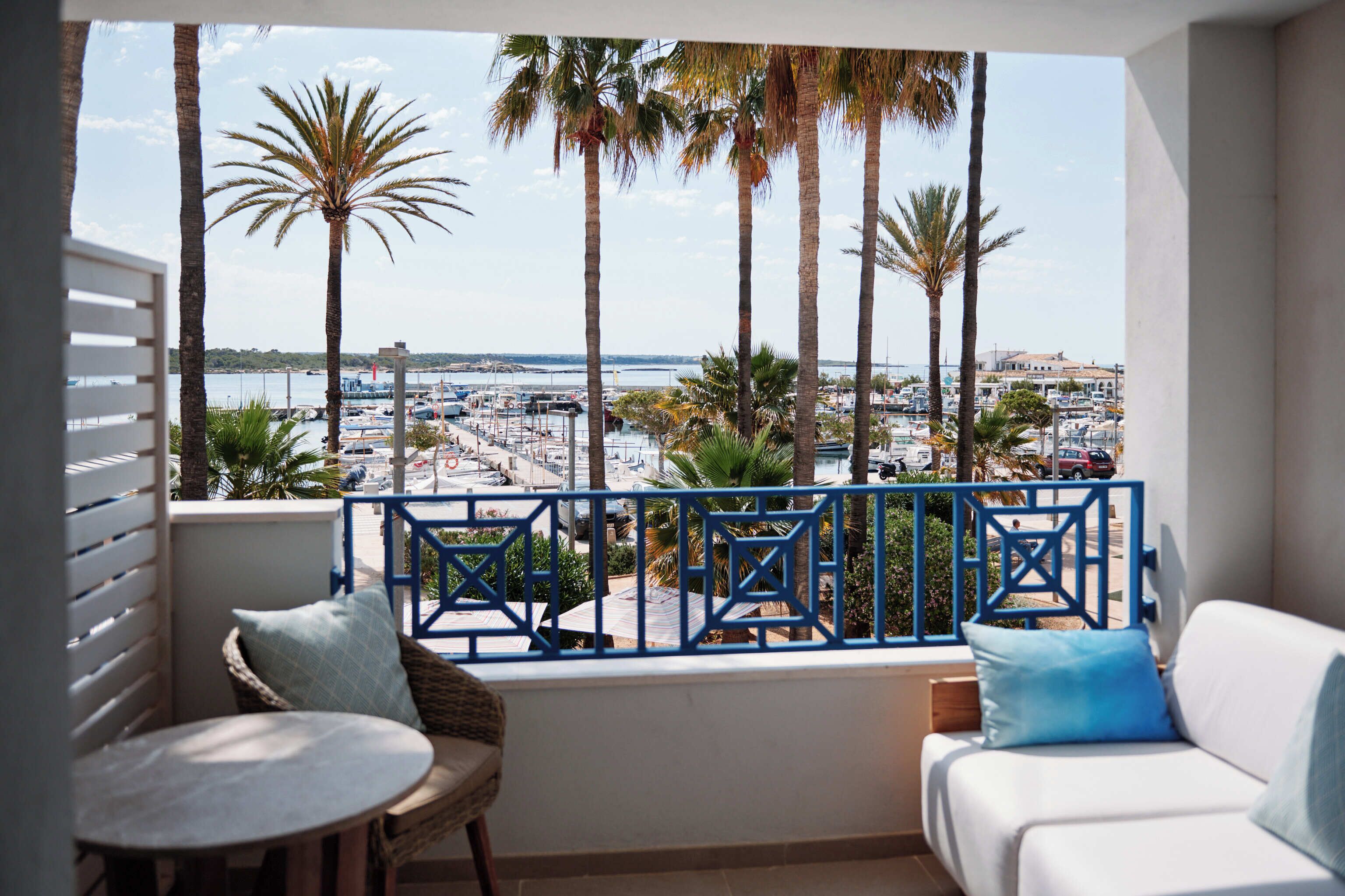A balcony with a white sofa and a small table, overlooking a marina with boats and palm trees.