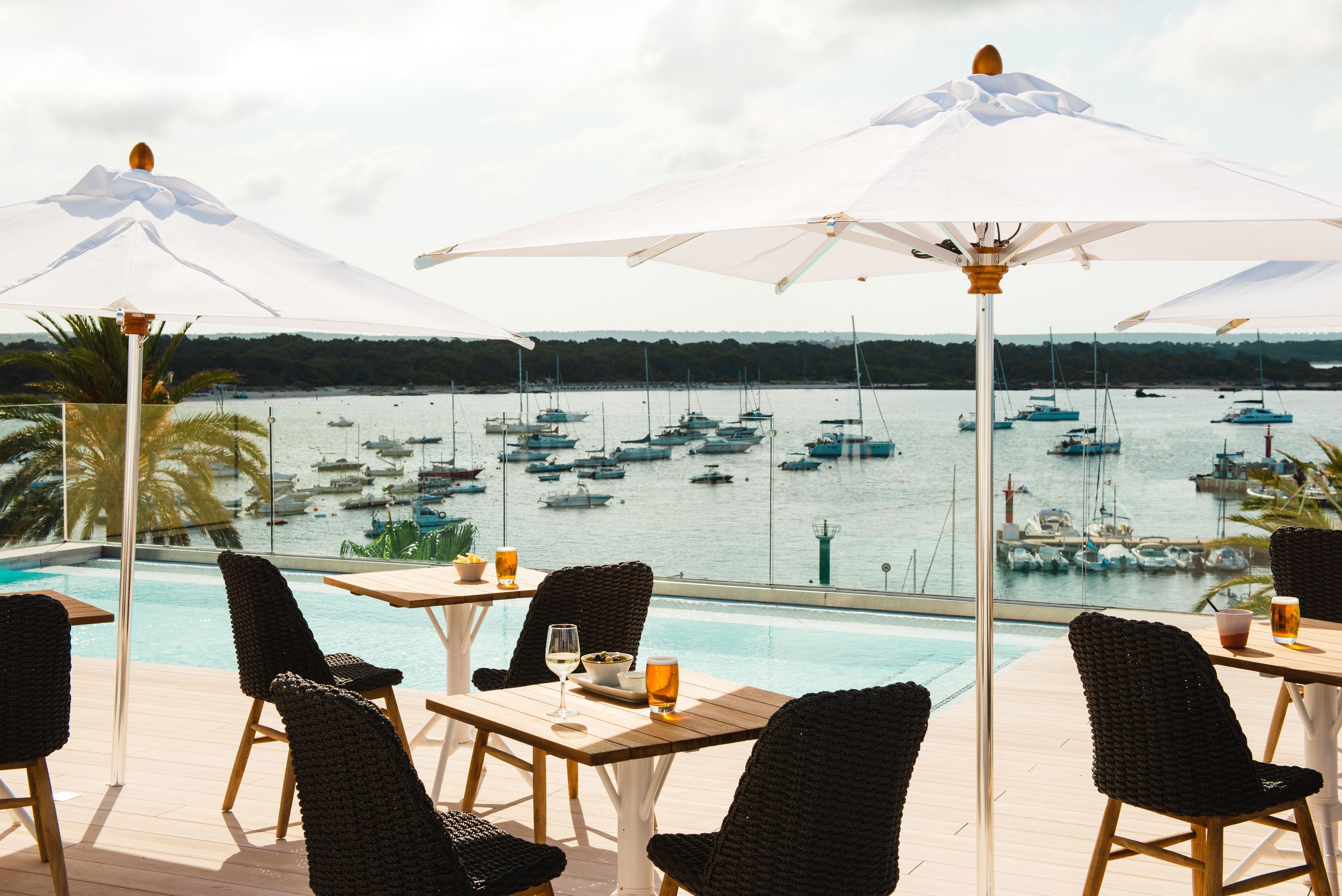 Zona de piscina con mobiliario exterior y sombrillas, con vistas a un puerto deportivo con barcos.