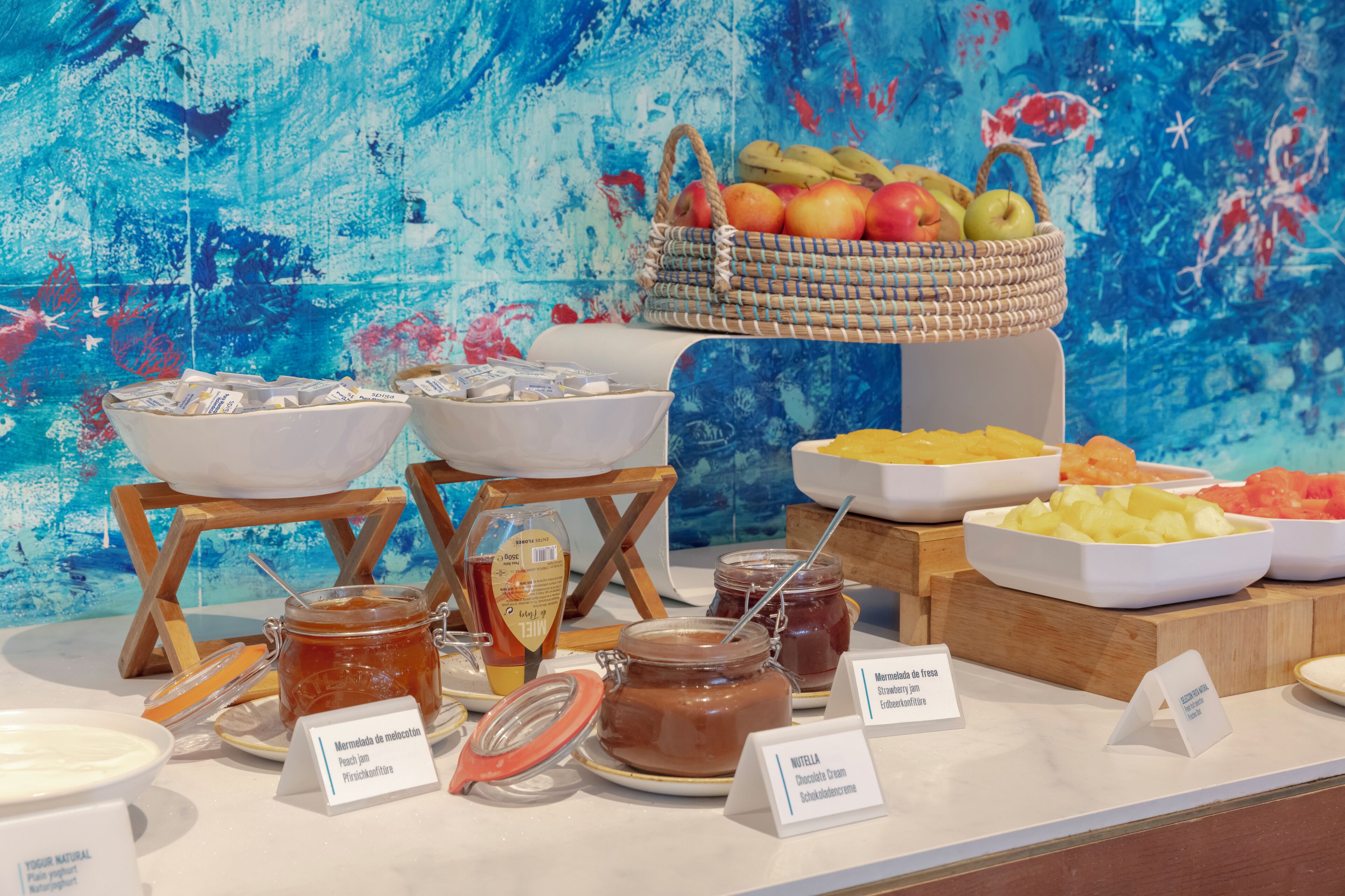 A buffet with various food items and condiments, including jars of honey and chocolate spread, and a basket of fruits.