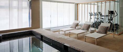 Un área de piscina interior moderna con cerramiento de vidrio, equipo de ejercicio y asientos.
