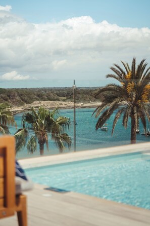 A pool area with a view of the sea, palm trees, and a distant shoreline. - Hotel Honucai (Ses Salines)
