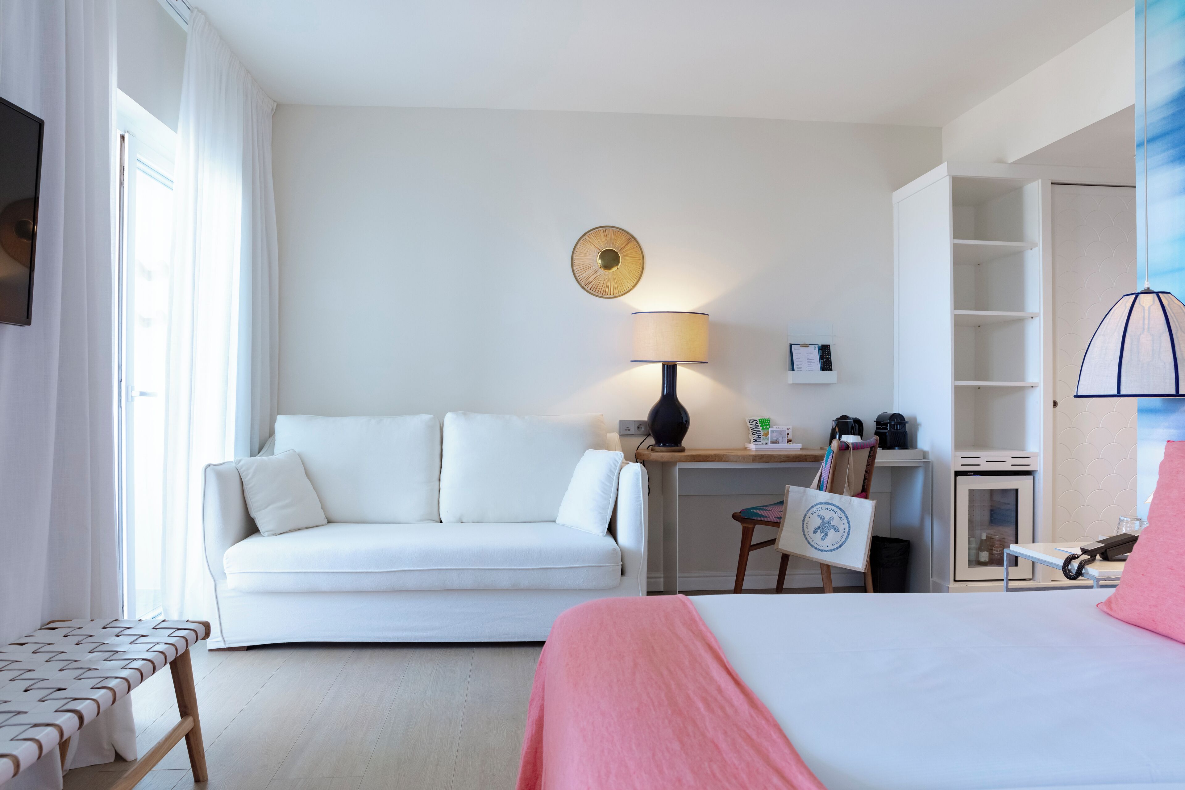A hotel room with a bed, a white sofa, a desk with a lamp, a small cabinet, and a chair.