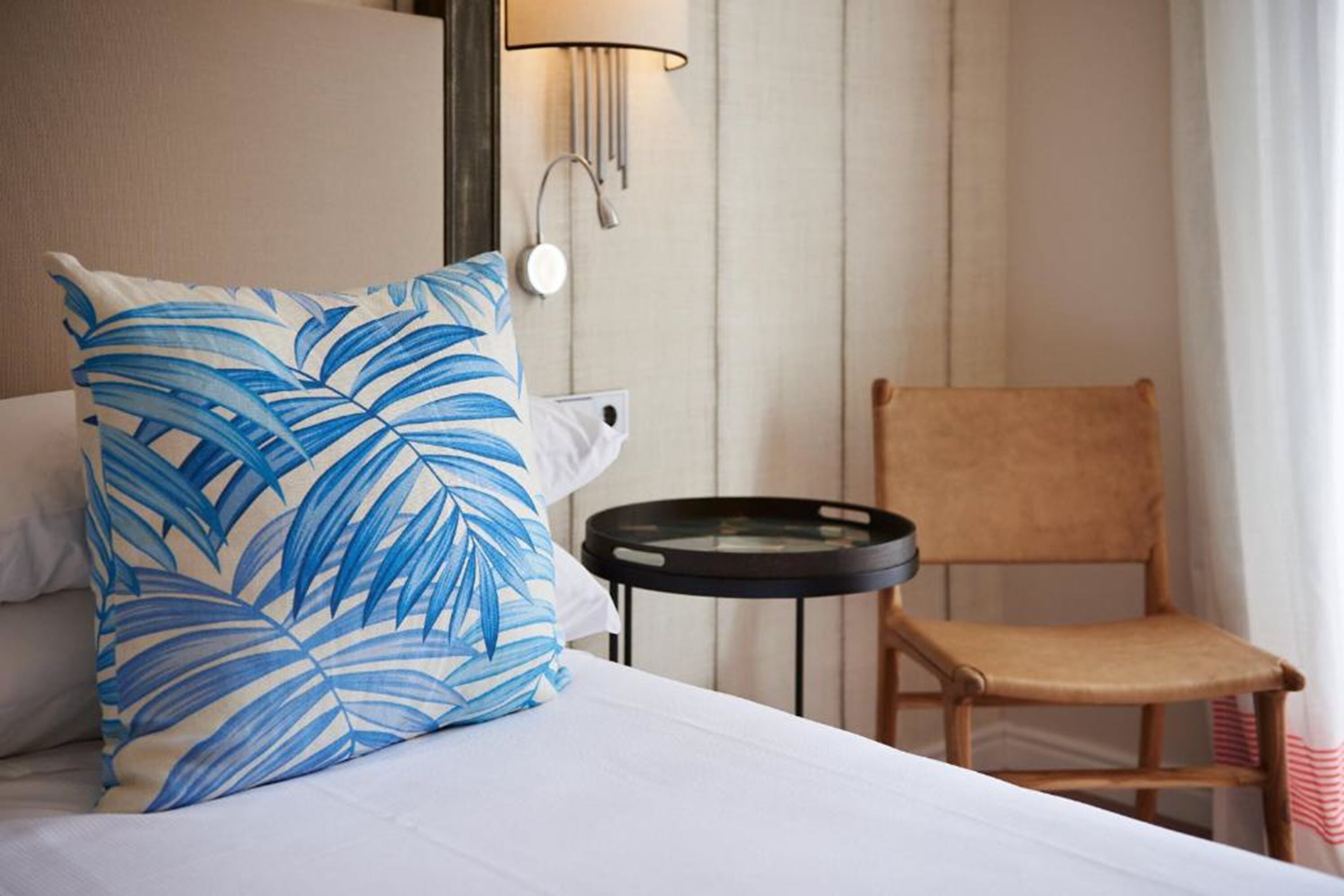 Habitación de hotel con cama, almohada estampada con motivos de hojas, una silla, una mesita y una lámpara de pared.
