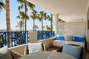 Ein Balkon mit Blick auf Palmen und das Meer, ein weißes Sofa mit blauen Kissen und ein Korbstuhl.